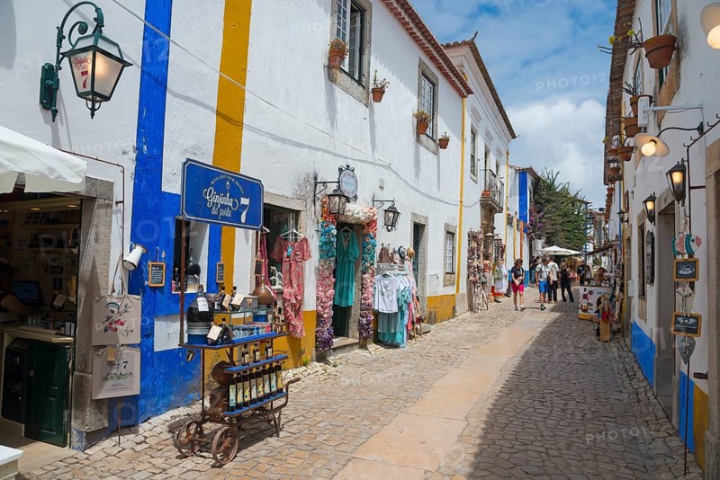 Narrow alley with souvenir shops and colourful clothes, blue and white facades and flowers, R. Direita, Óbidos, Obidos, Oeste, Centro, Estremadura, Portugal, Europe