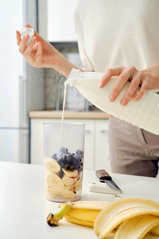 Unrecognizable woman pours milk from bottle into bowl of blender to make a smoothie with black currant