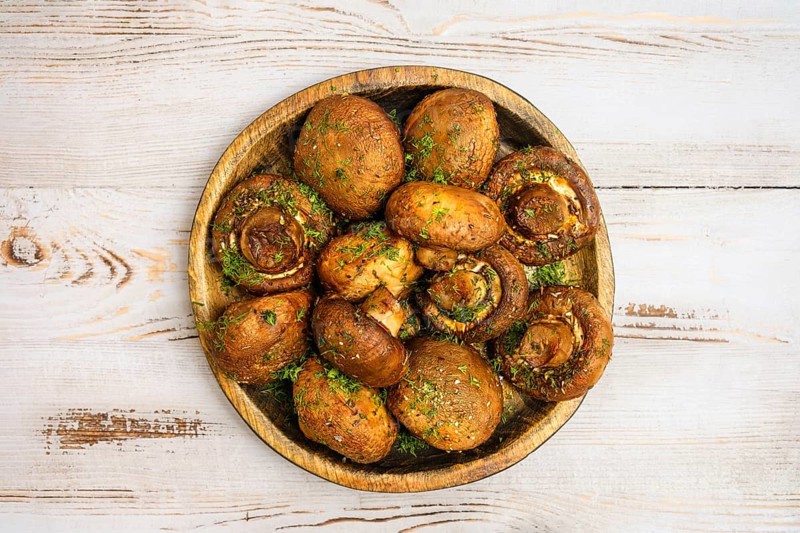 A rustic wooden plate with baked champignon mushrooms, each filled with herbs, sitting atop a light wooden table. The warm, inviting colors enhance the appetizing display.