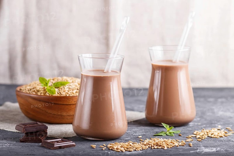 Organic non dairy oats chocolate milk in glass and wooden plate with oats seeds on a black concrete background. Vegan healthy food concept, close up, side view