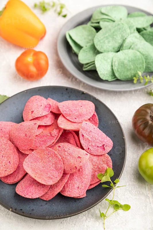 Green and red potato chips with herbs and tomatoes on gray concrete background. Side view, close up, selective focus