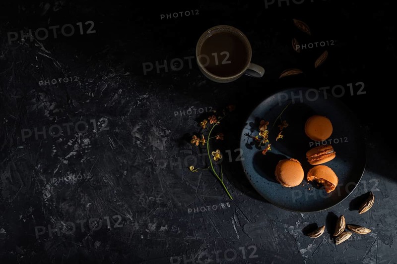 Orange macarons or macaroons cakes with cup of coffee on a black concrete background. Hard light, contrast, low key. Top view, flat lay, copy space
