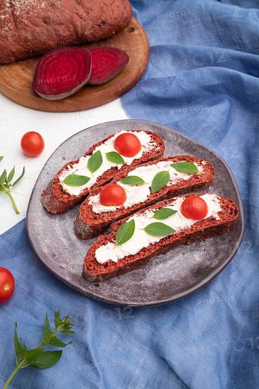 Red beet bread sandwiches with cream cheese and tomatoes on white concrete background and blue linen textile. Side view, close up