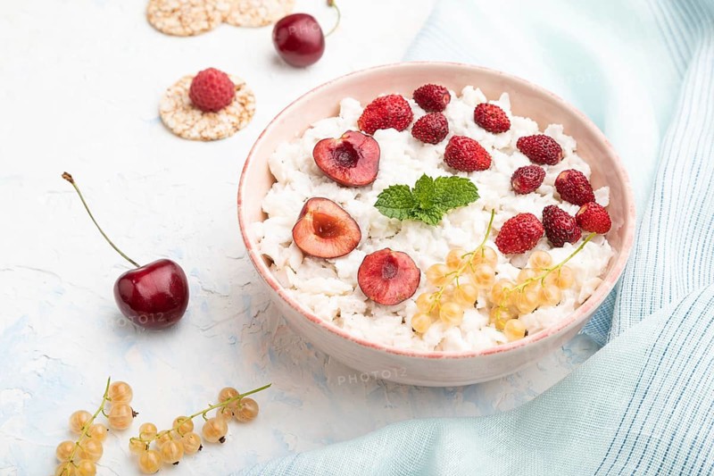 Rice flakes porridge with milk and strawberry in ceramic bowl on white concrete background and blue linen textile. Side view, close up