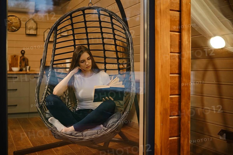 A young woman relaxes in the evening in a wooden house sitting in a hanging chair with a book