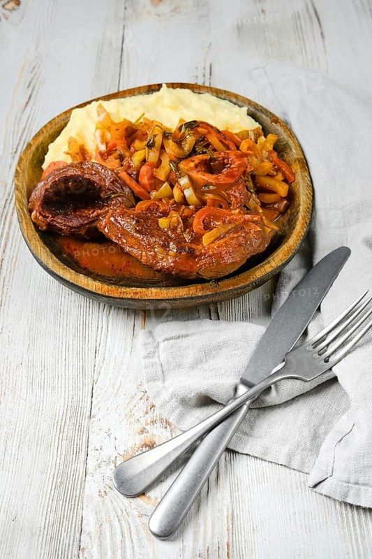 A wooden plate of osso buco served with mashed potatoes, cooked in a tomato and onion sauce with herbs. A silver knife and fork are placed beside a linen napkin on a white wooden table.