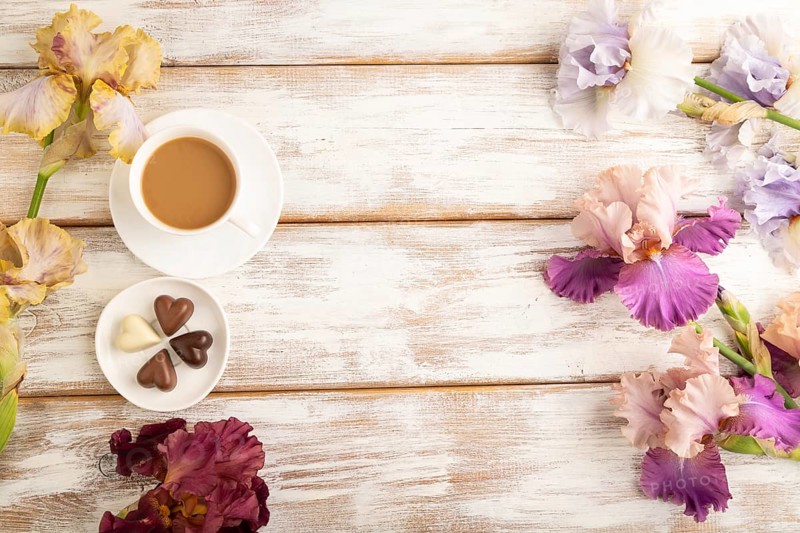 Cup of cioffee with chocolate candies, lilac and purple iris flowers on white wooden background. top view, flat lay, copy space, still life. Breakfast, morning, spring concept