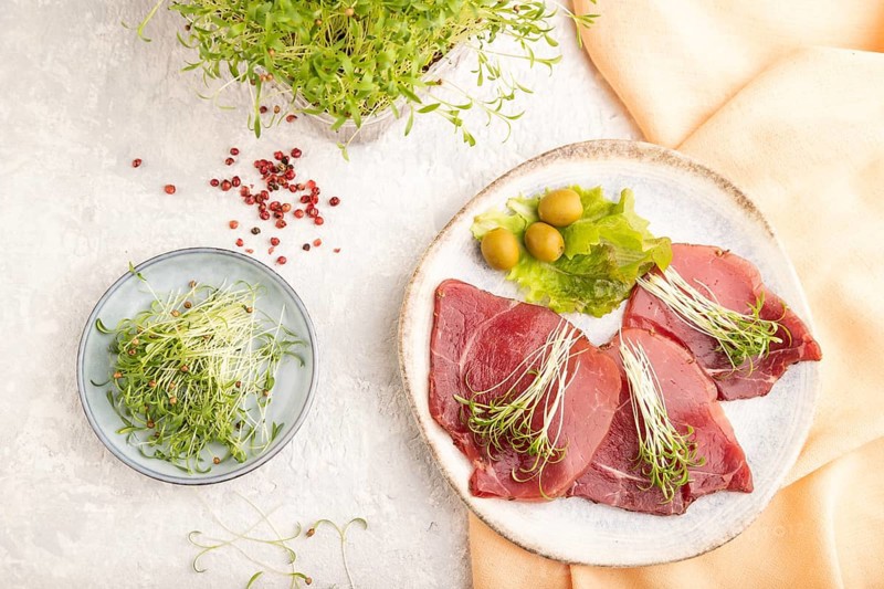 Slices of smoked salted meat with cilantro microgreen on gray concrete background and orange textile. Top view, flat lay, close up
