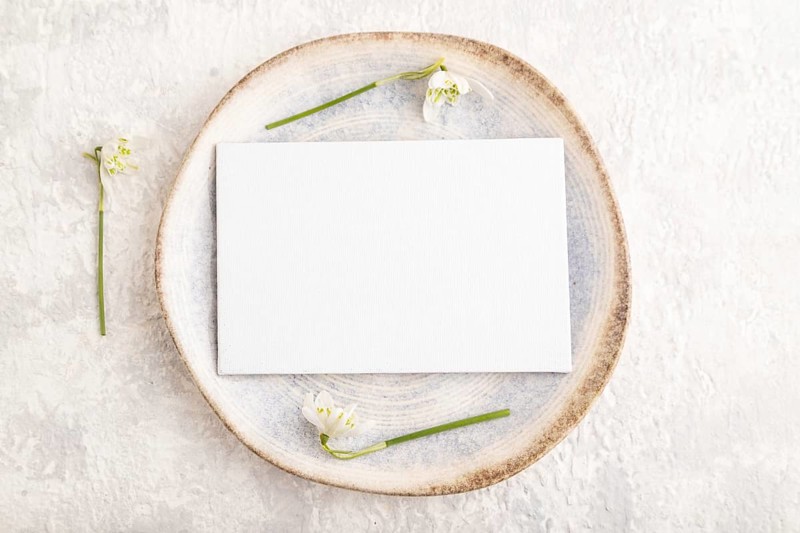 White paper invitation card, mockup with galanthus snowdrop flowers on ceramic plate and gray concrete background. Blank, flat lay, top view, still life, copy space, wedding invitation