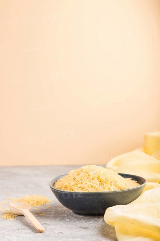 Blue ceramic bowl with raw golden rice and wooden spoon on a gray and orange background and yellow textile. Side view, selective focus, copy space