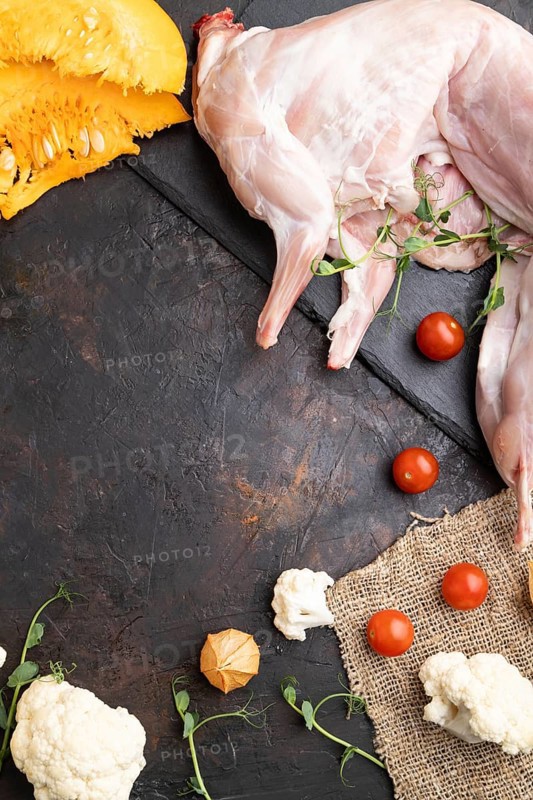 Whole raw rabbit with pumpkin, tomatoes, pea sprouts on a black concrete background and linen textile. Top view, flat lay, copy space