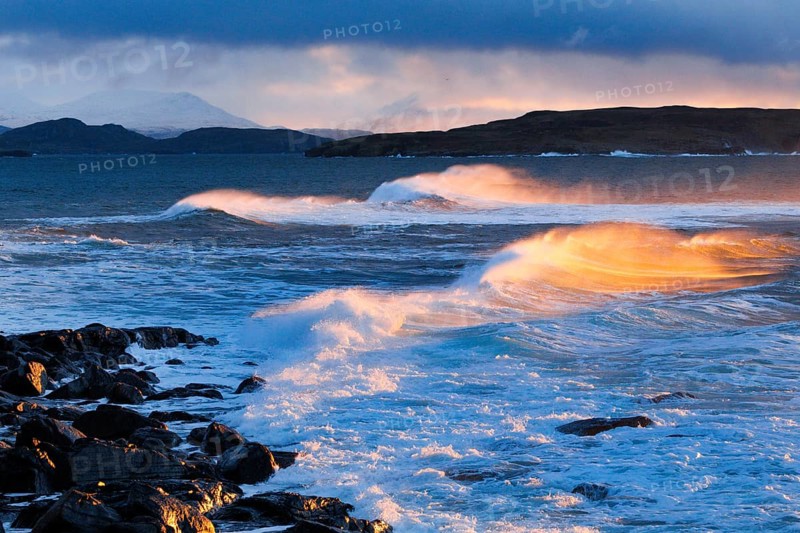 Large waves crash in a winter storm and the swirling spray is illuminated by the warm light of the evening sun, Summer Isles in the background, at Reiff on the north-west coast of Scotland