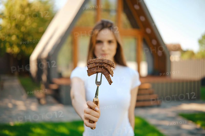 Defocused young woman stretching forward a barbecue fork with slices of steak in her hand