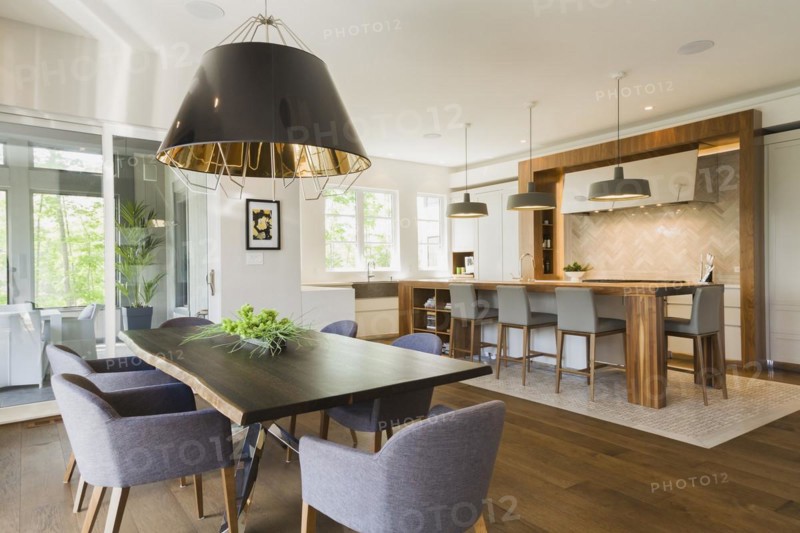 Chrome and walnut wood table with charcoal linen armchairs, large black and gold industrial style pendant lighting fixture and walnut wood island with grey leather and wooden high chairs on travertine mosaic floor surrounded by engineered hickory wood floorboards in dining room kitchen area inside a luxurious contemporary bungalow style home, Quebec, Canada, North America