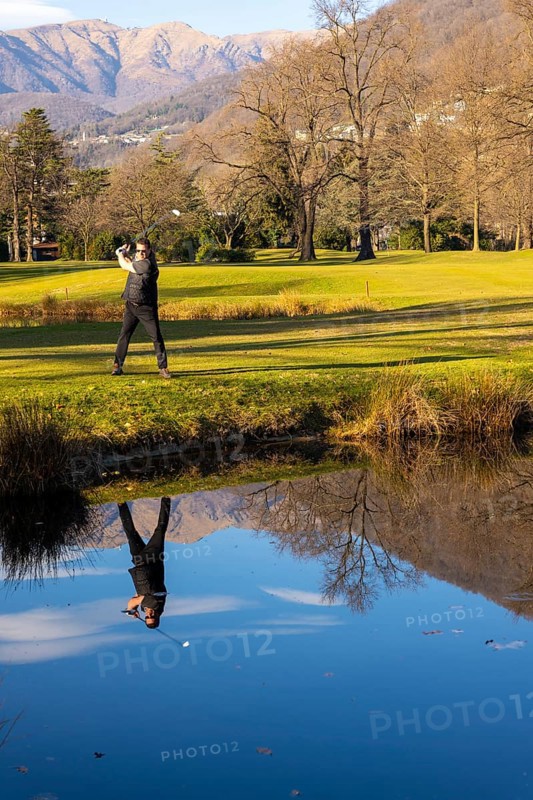 Male Golfer Reflected in a Water Pond and Hitting the Golf Ball on Fairway on Golf Course with Mountain in a Sunny Day in Switzerland