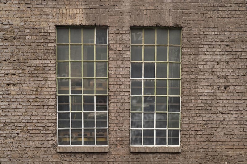 Two large industrial windows with small panes and metal frames are set in an old brick wall, revealing pallets inside the building