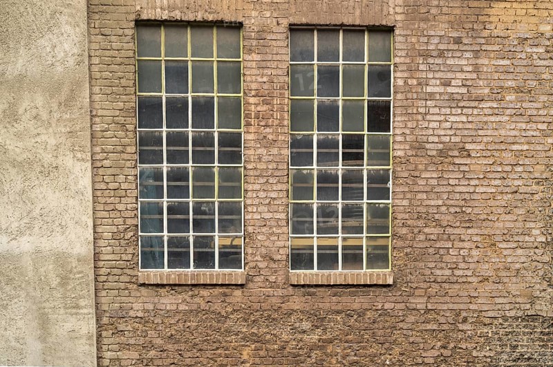 Two large industrial windows with small panes and metal bars are set in an old brick wall, showing signs of age and wear