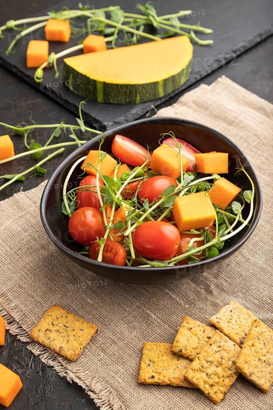 Vegetarian vegetable salad of tomatoes, pumpkin, microgreen pea sprouts on black concrete background and linen textile. Side view, close up