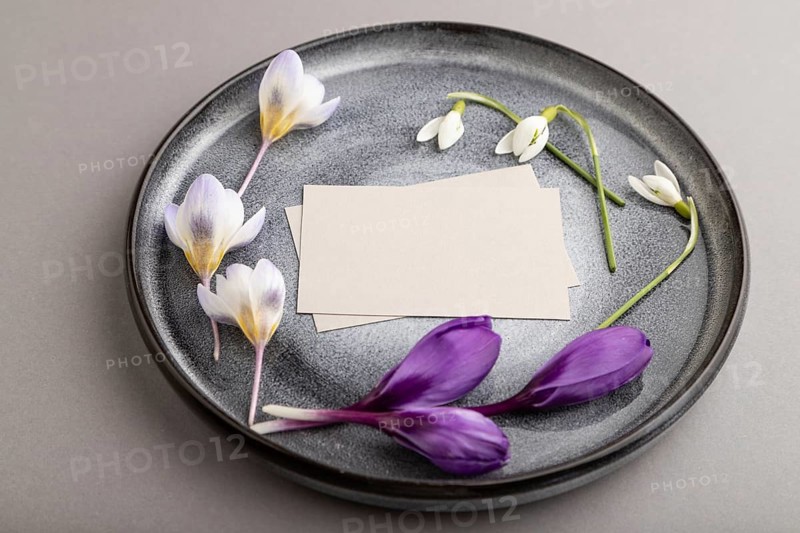 Gray paper invitation card, mockup with crocus and galanthus flowers on ceramic plate and gray background. Blank, side view, still life, copy space, wedding invitation