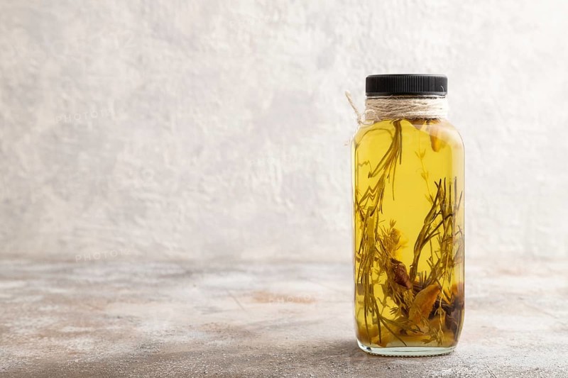 Sunflower oil in a glass jar with various herbs and spices, lavender, sesame, rosemary on a brown concrete background. Side view, copy space