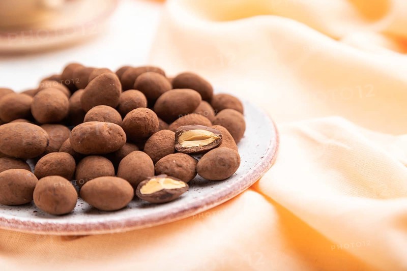 Almond in chocolate dragees on ceramic plate and a cup of coffee on white concrete background and orange linen textile. Side view, close up, selective focus