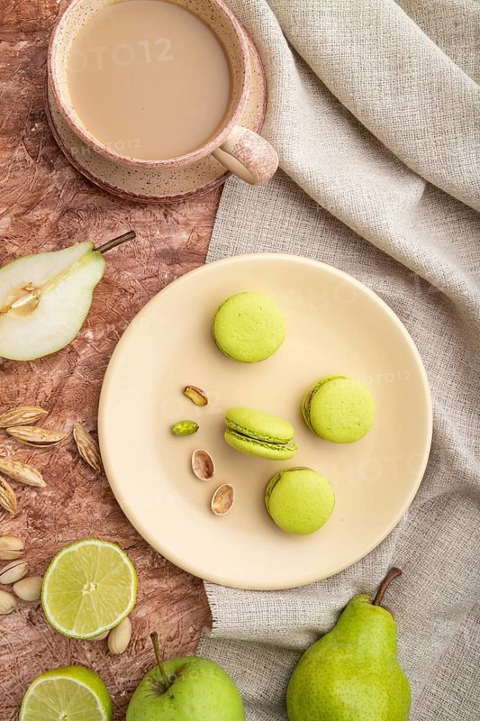 Green macarons or macaroons cakes with cup of coffee on a brown concrete background and linen textile. Top view, flat lay, close up