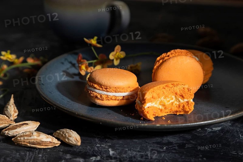 Orange macarons or macaroons cakes with cup of coffee on a black concrete background. Hard light, contrast, low key. Side view, close up