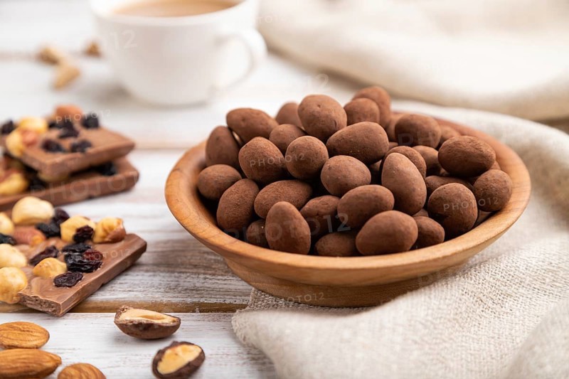 Almond in chocolate dragees in wooden plate and a cup of coffee on white wooden background and linen textile. Side view, close up, selective focus