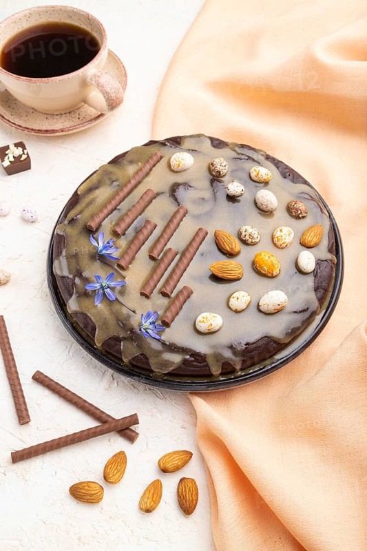 Homemade chocolate brownie cake with caramel cream and almonds with cup of coffee on a white concrete background and orange textile. Side view, close up