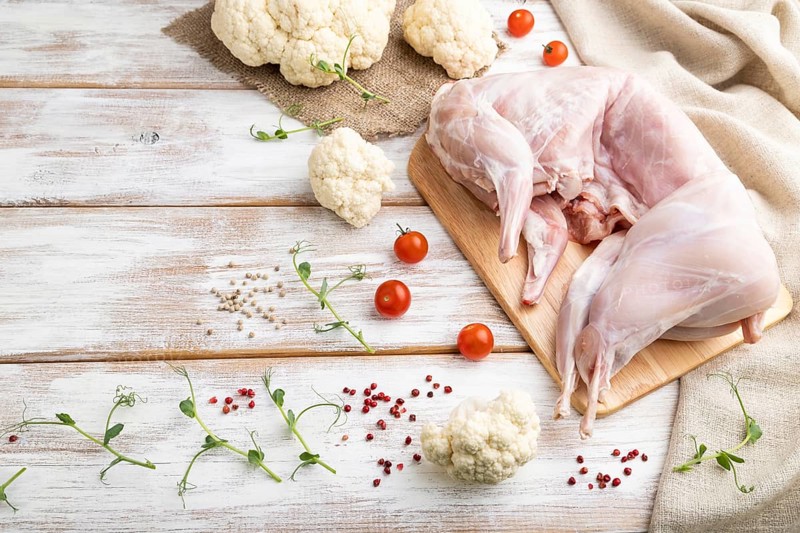 Whole raw rabbit with cauliflower, tomatoes and spices on a white wooden background and linen textile. Side view, copy space