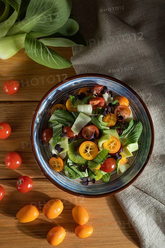 Vegetarian salad of pac choi cabbage, kiwi, tomatoes, kumquat, microgreen sprouts on a wooden background and linen textile. Hard light, contrast. Top view, flat lay, copy space