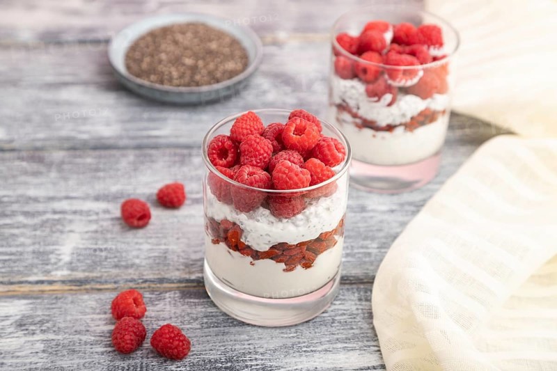 Yogurt with raspberry, goji berries and chia seeds in glass on gray wooden background and linen textile. Side view, close up, selective focus
