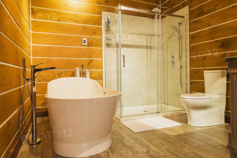 Freestanding bathtub with high tank toilet and clear glass shower stall in en suite with imitation wood ceramic tile flooring inside a milled Eastern white pine timber and flat log profile home, Quebec, Canada, North America