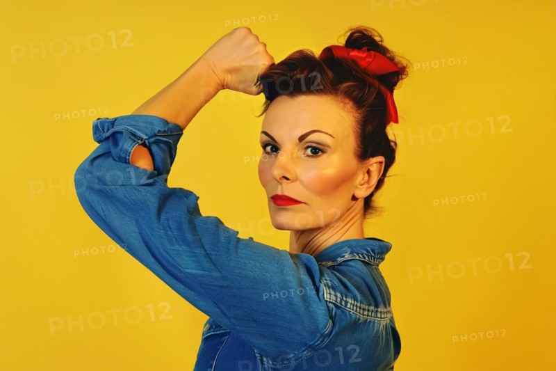 Woman with retro hairstyle and blue blouse raising her hand in fist in front of yellow studio background. Feminism concept. KI generiert, generiert, AI generated
