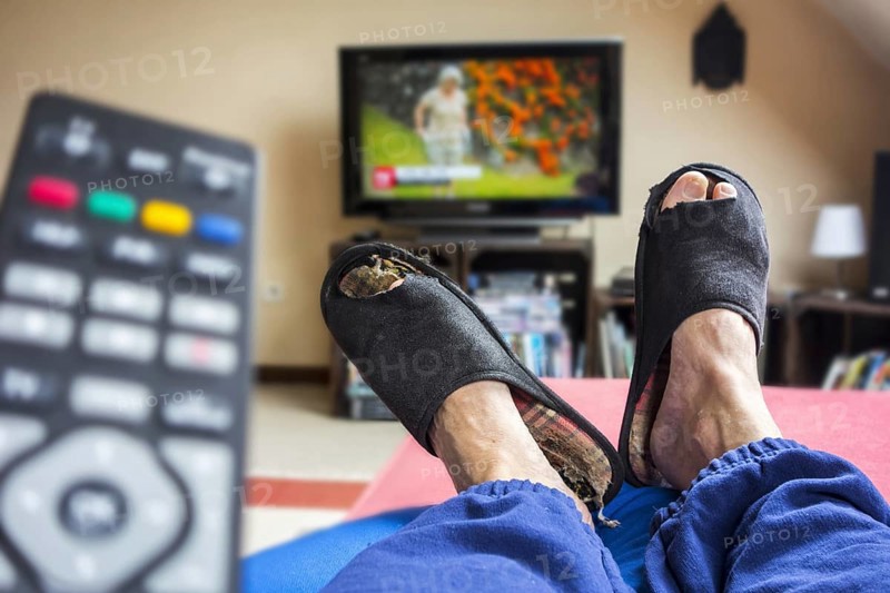 Remote control and couch potato, lazy man in comfy chair wearing worn slippers with big toes sticking through and watching television in living room