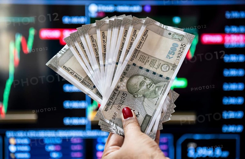 A person holding Indian 500 rupee currency bank note against share market graph on a display