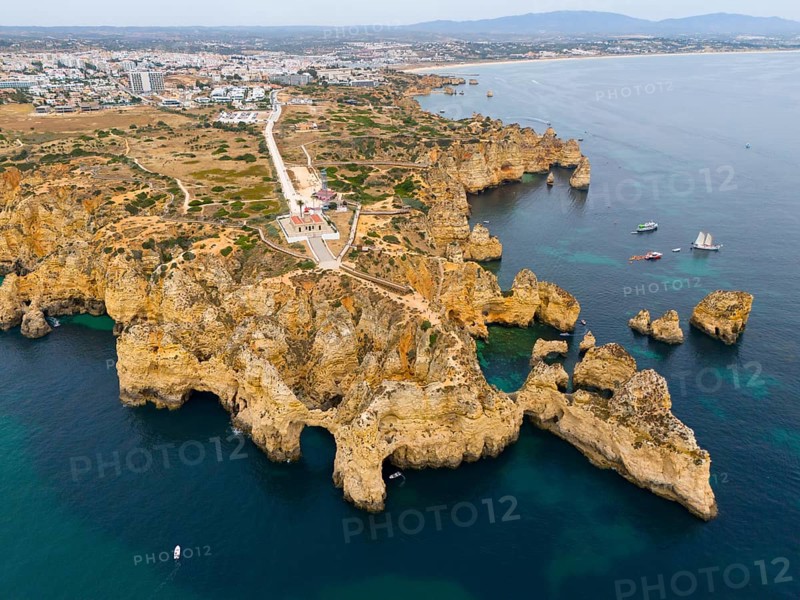 Steep cliffs and rocky coastline with blue sea, beyond a town and distant mountains, aerial view, Ponta da Piedade, point of mercy, Lagos, Algarve, Portugal, Europe