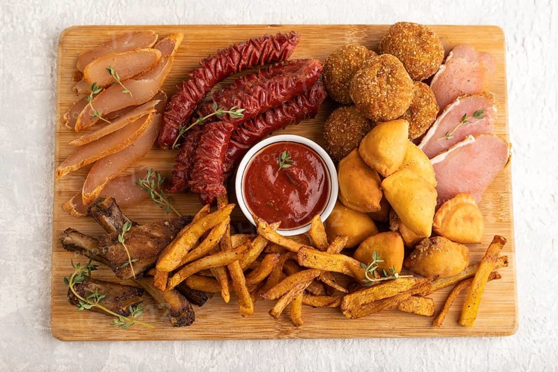 Set of snacks: sausages, fried potatoes, meat balls, dumplings, basturma on a cutting board on a gray concrete background. Top view, flat lay, close up