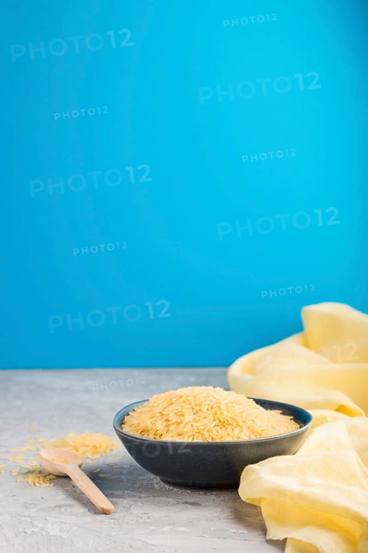 Blue ceramic bowl with raw golden rice and wooden spoon on a gray and blue background and yellow textile. Side view, selective focus, copy space