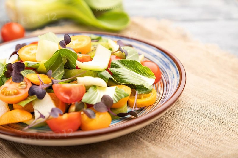 Vegetarian salad of pac choi cabbage, kiwi, tomatoes, kumquat, microgreen sprouts on gray wooden background and linen textile. Side view, close up, selective focus