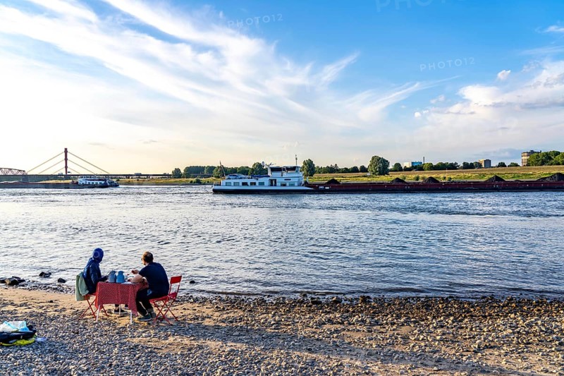 The Rhine at Duisburg-Baerl, man and woman having a picnic on the Rhine beach, at low tide, on the other side of the Rhine the Thyssenkrupp Steel steelworks and Duisburg-Beeckerwerth, cargo ships, coal freighters, Duisburg, North Rhine-Westphalia, Germany, Europe