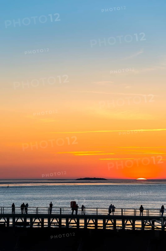 Silhouettes of several people on the pier free canal, couple and single persons watch from a jetty the picturesque, golden, breathtakingly beautiful sunset over the sea, island Langlütjen II far away on the horizon, sun almost completely sunk, crescent, orange and blue glowing cloudless sky, wide sea in soft colours, calm atmosphere, mouth of the Weser into the North Sea, Outer Weser, Bremerhaven, Land Bremen, Germany, Europe