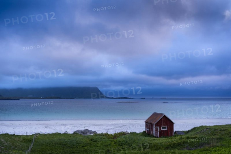 A red wooden hut on the sandy beach of Ramberg (Rambergstranda), sea and mountains. At night at the time of the midnight sun. Clouds in the sky. Early summer. Ramberg, Flakstadoya, Lofoten, Norway, Europe