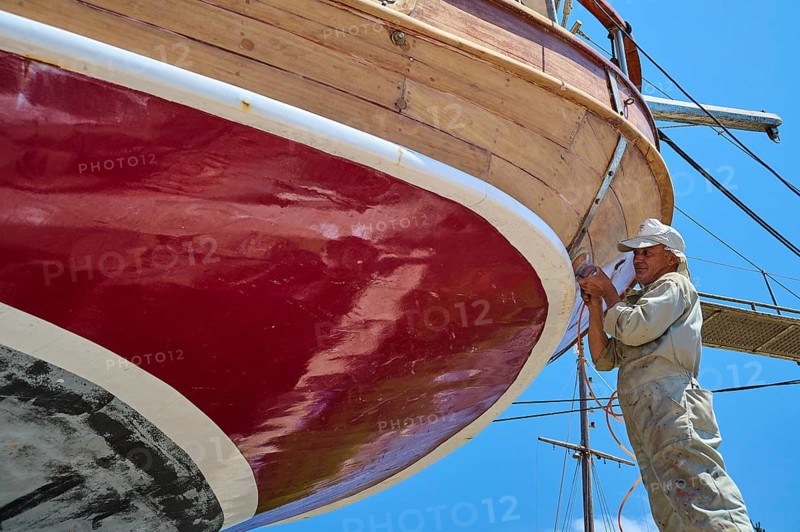 A worker is working on the red hull of a sailing ship, Patmos Marine, boatyard, Diakofti, Patmos, Dodecanese, Greek Islands, Greece, Europe