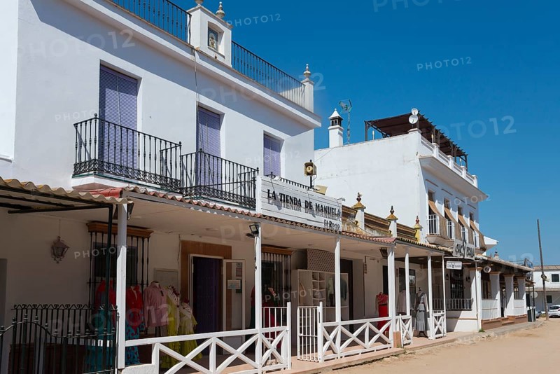 Eine sonnige Straßenszene mit weißen Gebäuden und Geschäftsfassaden im spanischen Stil unter blauem Himmel, Wallfahrtsort, El Rocío, Rocío, Rocio, Huelva, Andalusien, Spanien