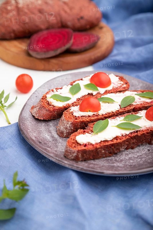 Red beet bread sandwiches with cream cheese and tomatoes on white concrete background and blue linen textile. Side view, selective focus, close up