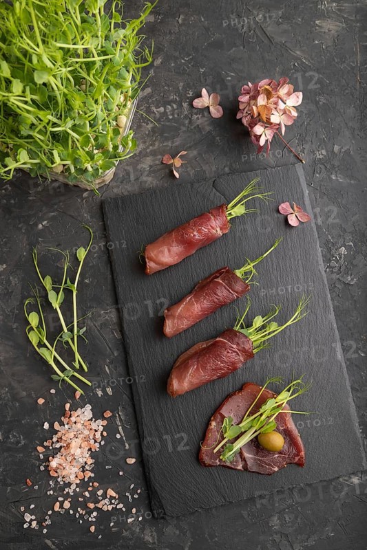 Slices of smoked salted meat with green pea microgreen on black concrete background. Top view, flat lay, close up