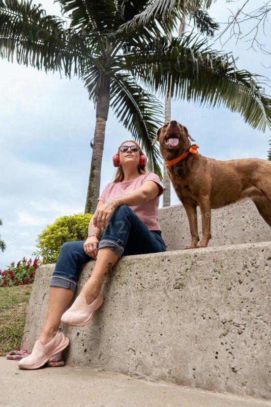 Middle-aged woman seated outdoors wearing red headphones and sunglasses, enjoying the sunny day with her pet