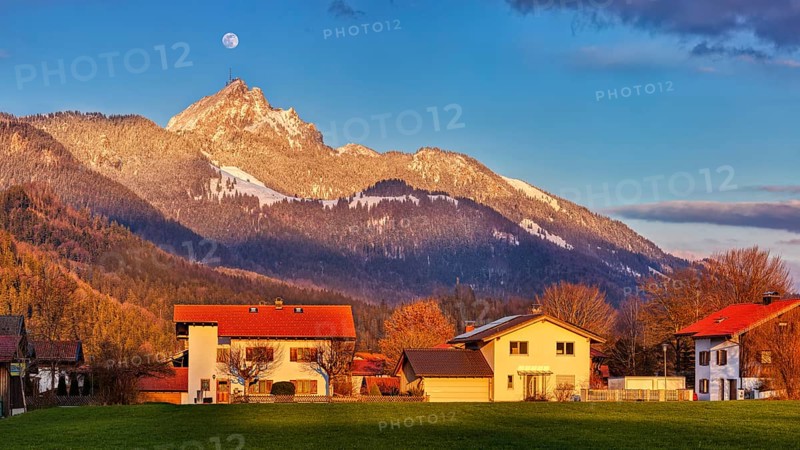 Sunset over an alpine scene with houses in the foreground, snow-covered mountain in the background and visible moon, Wendelstein
