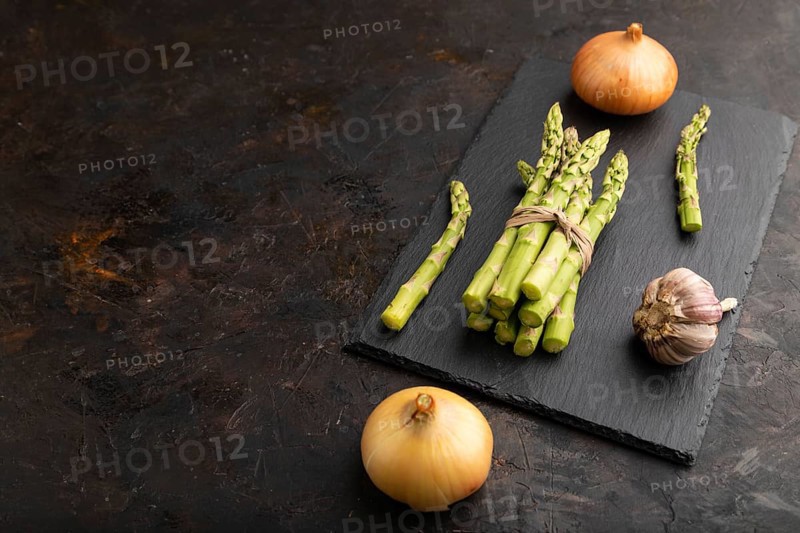 Bunch of fresh green asparagus, garlic, onion on slate board on black concrete background. Side view, copy space. harvest, healthy, vegan food, concept